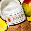 A jar of RootMask- Vigor Roots' reparative hair mask is posed with fresh coconut & mango- some of the food-grade, all natural ingredients featured in the luxurious formula.