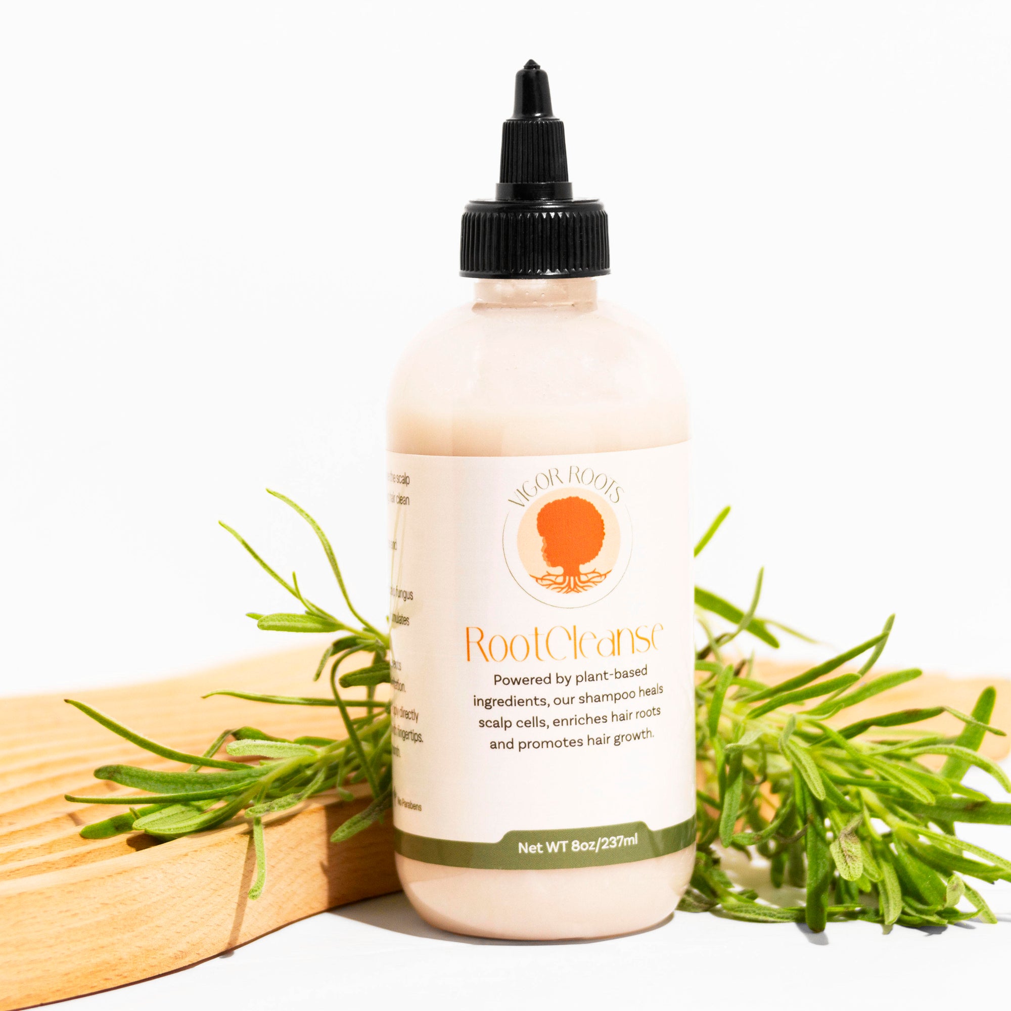 A bottle of Vigor Roots RootCleanse Shampoo is posed with a fresh sprig of rosemary- one of the food-grade, all-natural ingredients included in the luxurious formula.